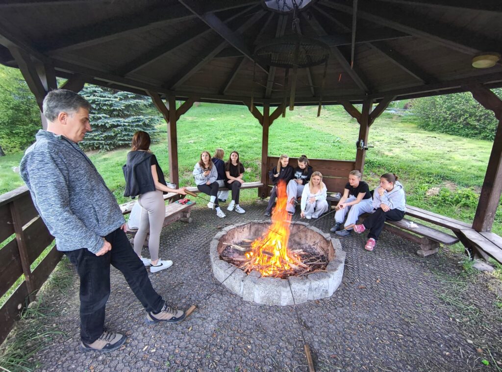 uczennice siedzące przy ognisku, obok stojący pedagog szkolny